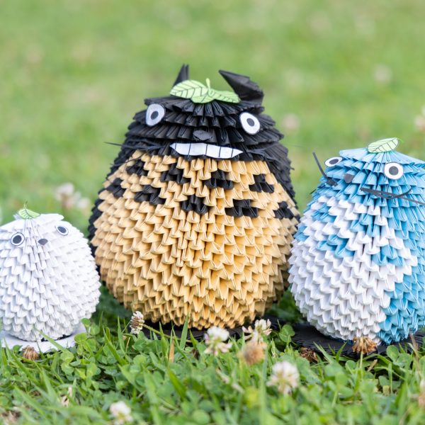 芝生の上に並んだブロック折り紙の立体作品3体。小さく折った紙パーツを組み上げて作られている。中央に黒と黄土色の大トトロ、右に水色と白の中トトロ、左に白い小トトロ。3体とも白い丸い目を持ち、頭に緑色の葉を載せている。大トトロは白い歯を見せて笑い、胸に黒い模様がある。