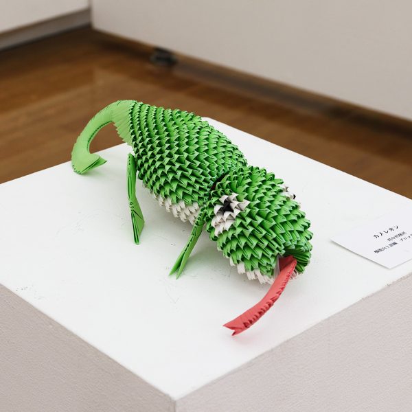 緑色の三角パーツを鱗のように積み上げた立体折り紙のカメレオン。体を丸め、四本の脚と巻いた尾をもち、口から赤く細長い舌を伸ばす。腹部は白い。