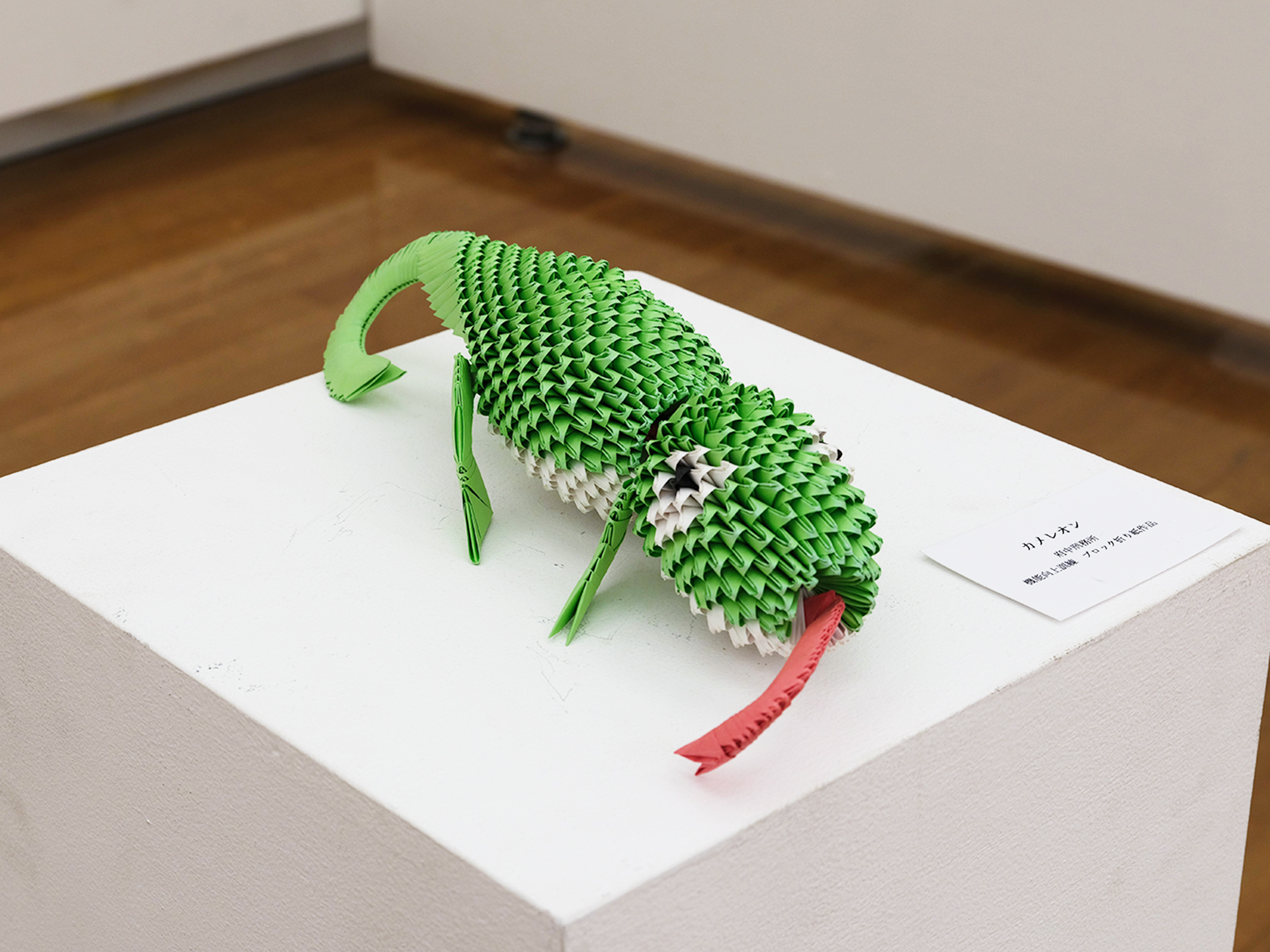 緑色の三角パーツを鱗のように積み上げた立体折り紙のカメレオン。体を丸め、四本の脚と巻いた尾をもち、口から赤く細長い舌を伸ばす。腹部は白い。