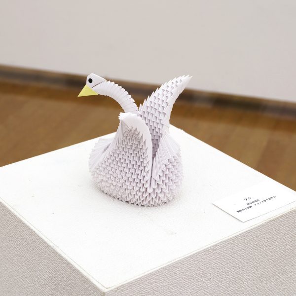 白（淡い藤色）の小さな三角パーツを多数組み上げた立体折り紙。長い首をゆるやかにもたげ、黄色いくちばしと黒い目をもつ白鳥のような鳥を表す。左右に翼を広げている。