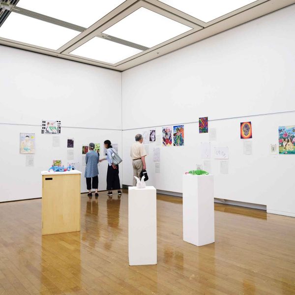 府中市美術館市民ギャラリーの展示風景。白い壁面に応募作品の絵画が並び、来場者が鑑賞している。中央奥の壁面に、少年漫画のキャラクターを描いたペン画《ジャンプヒーローズ》が展示されている。