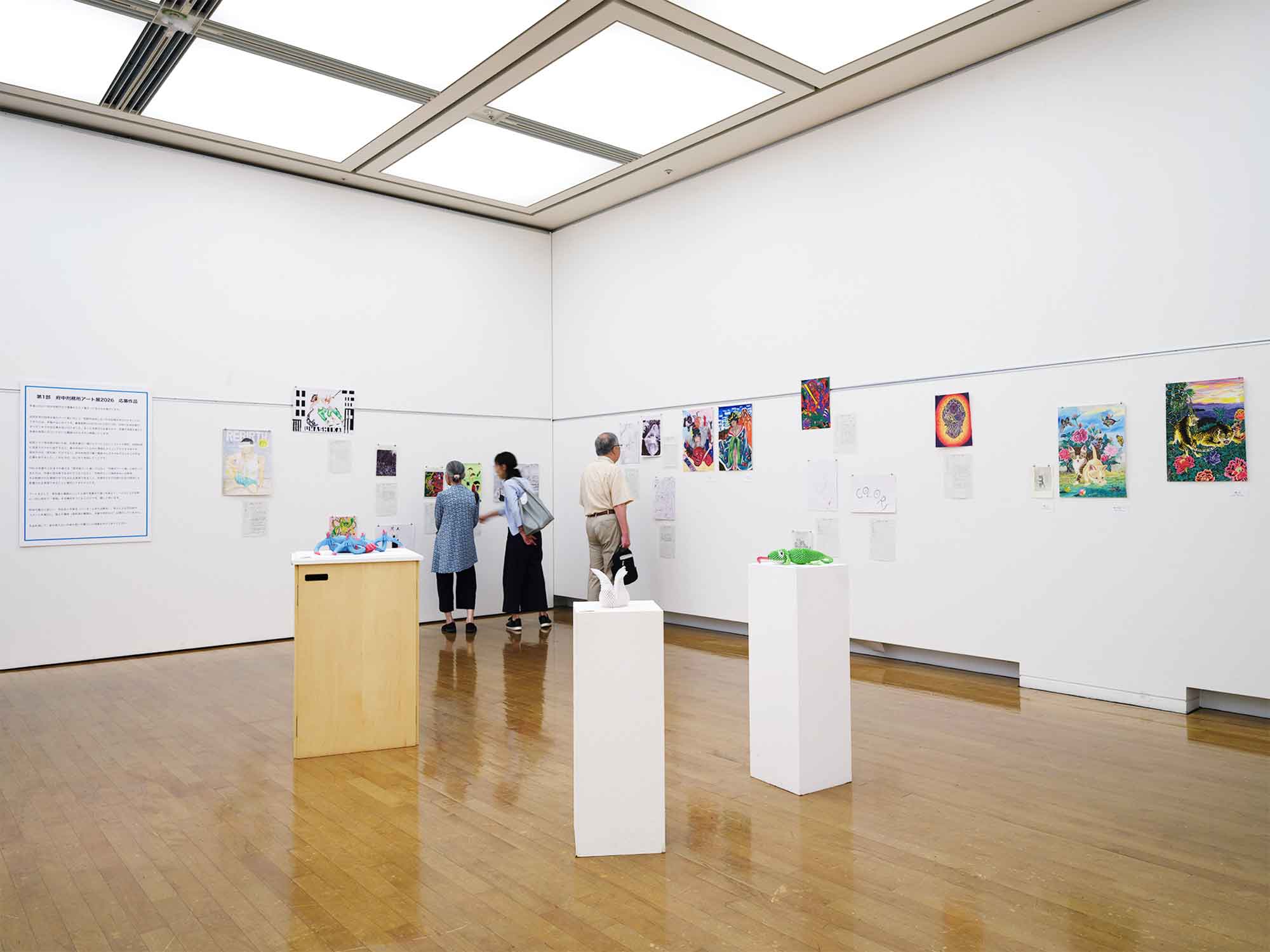 府中市美術館市民ギャラリーの展示風景。白い壁面に応募作品の絵画が並び、来場者が鑑賞している。中央奥の壁面に、少年漫画のキャラクターを描いたペン画《ジャンプヒーローズ》が展示されている。