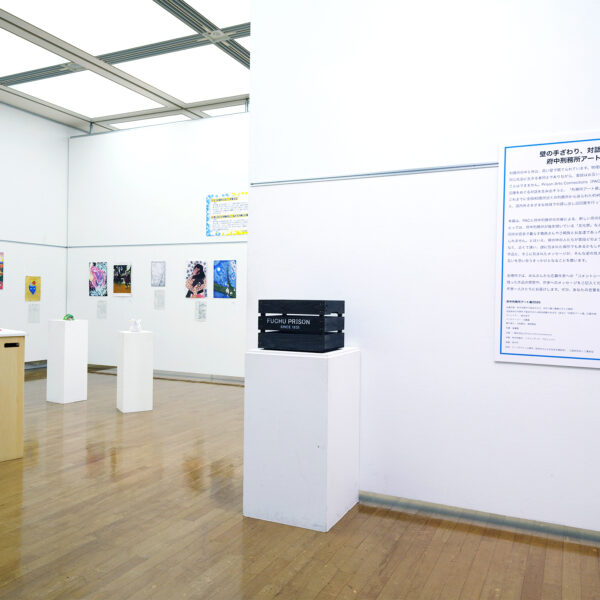 府中刑務所アート展2026の会場入口付近。壁面に絵画・書・折り紙作品が並び、右手に展覧会概要パネルと「FUCHU PRISON」と書かれた木箱の展示台