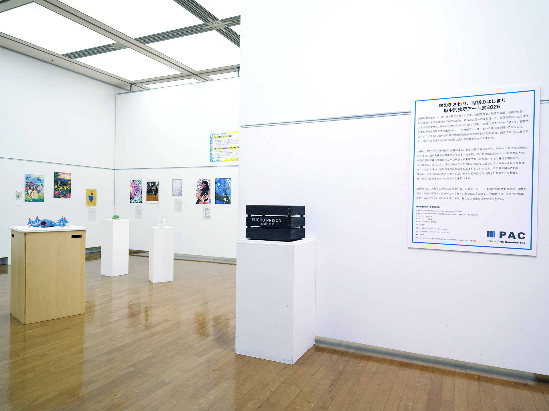 府中刑務所アート展2026の会場入口付近。壁面に絵画・書・折り紙作品が並び、右手に展覧会概要パネルと「FUCHU PRISON」と書かれた木箱の展示台