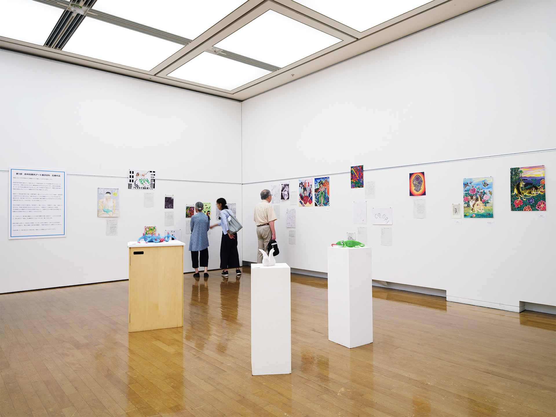 第1部の展示風景。壁一面に応募作品の絵画が並び、来場者3名が作品を鑑賞している様子